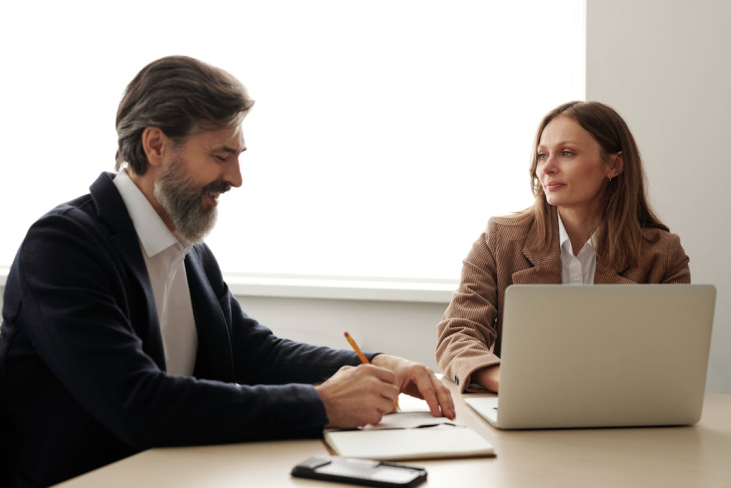 An image of a man writing in a notebook and a woman working on a laptop
